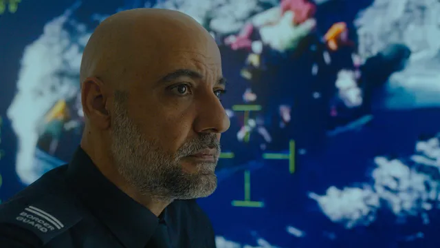 Man met baard zonder haar op hoofd, met overhemd waarop de schouders 'border guard' staat, kijkt serieus naar rechts. Op de achtergrond zien we dronebeelden van een boot met vluchtelingen op zee.