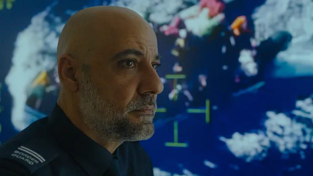 Man met baard zonder haar op hoofd, met overhemd waarop de schouders 'border guard' staat, kijkt serieus naar rechts. Op de achtergrond zien we dronebeelden van een boot met vluchtelingen op zee.