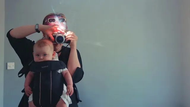 Een vrouw met een camera voor haar gezicht draagt een baby in een draagzak op haar buik.