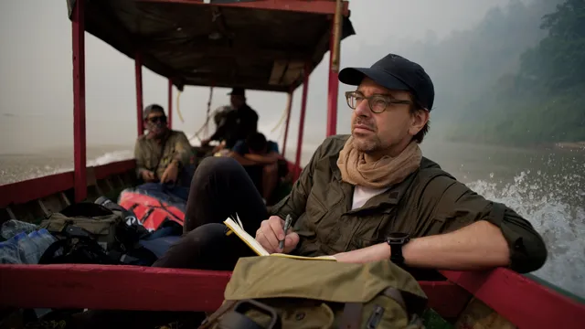 Ruben Terlou, een witte man met bril, pet en sjaal, zit met een notitieboek op een houten boot op een rivier, met junglegebied op de achtergrond. Op de boot zitten nog drie mensen, deels onscherp in beeld, omringd door bagage.