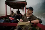 Ruben Terlou, een witte man met bril, pet en sjaal, zit met een notitieboek op een houten boot op een rivier, met junglegebied op de achtergrond. Op de boot zitten nog drie mensen, deels onscherp in beeld, omringd door bagage.