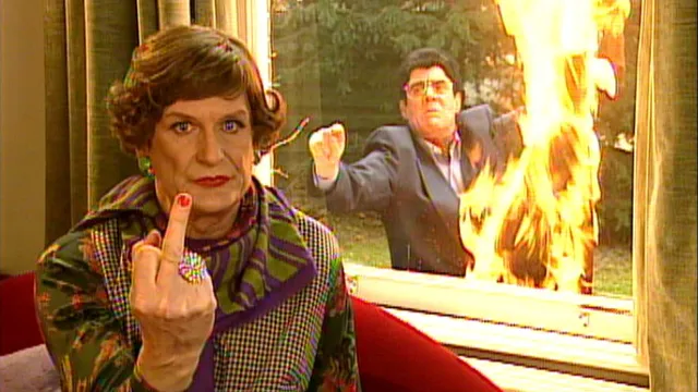 Een wit persoon in travestie, Wim de Bie, zit op een bank en steekt zijn middelvinger op naar de camera. Op de achtergrond staat een witte man in pak, Kees van Kooten, buiten voor het raam, terwijl er vlak naast hem grote vlammen omhoog slaan.