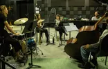 Een jazzensemble tijdens een repetitie in een studio met geluidsisolerende wanden. In het midden zit saxofoniste Kika Sprangers lachend in gesprek met een zangeres. Ze worden omringd door een drummer aan de linkerkant, en een pianist en contrabassist aan de rechterkant. De ruimte staat vol muziekstandaarden, wat een actieve en creatieve werksfeer weergeeft.
