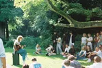 Een foto in analoge stijl van een intiem akoestisch optreden in de buitenlucht. Links zit muzikant Maxime met een gitaar op een zwarte versterker op het gras. Rechts luistert een groep jonge mensen, zittend en staand op het gazon onder een grote boom, naar het optreden.