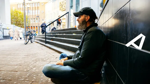 Man zit te bedelen tegen een muur met een bekertje in zijn hand.