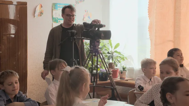 Witte man filmt met een videocamera op statief in een klaslokaal, terwijl kinderen aan tafels zitten bij een raam met planten op de vensterbank.