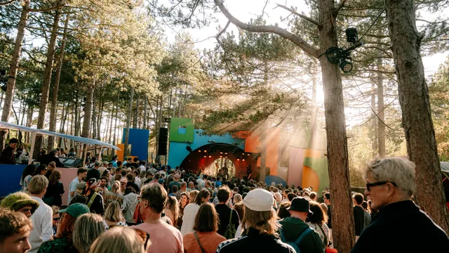 Mensen zitten voor een podium tussen de bomen, de zon schijnt door de takken