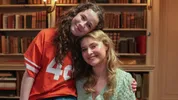 Roos draagt een rood ijshockey shirt en zit op schoot bij Vajèn, die een turqoise linnen blouse draagt. Ze zitten in een boekenkamer
