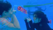 Twee meisjes met zwart haar gebaren naar elkaar onder water. Tussen hen in een kindertekening van een octopus