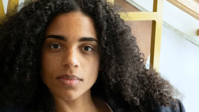 Een vrouw met zwarte krullen en in een witte top kijkt in de camera.