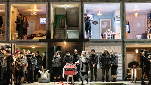 Verschillende mensen staan rondom een kist. Op de achtergrond zie je een dwarsdoorsnede van een flatgebouw. Het is een scene uit de opera Boris Godoenov