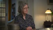Een dame met grijs haar en bril kijkt opzij in een woonkamer