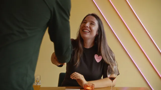 Vrouw in zwart shirt met papieren hartje opgespeld steekt geeft man een hand. Voor haar staat een kaarsje en een glas wijn.