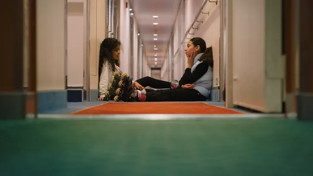 Twee kinderen zitten tegenover elkaar in de gang van een cruiseship, het meisje op rechts draagt skeelers aan haar voeten.