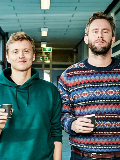 Twee witte mannen, Nicolaas Veul links en Tim den Besten rechts, lopen door een schoolgang; de een draagt een groene hoodie en de ander een kleurrijke trui en houdt papieren vast. Op de achtergrond zitten vier jonge mensen op een bank, onder wie een zwarte vrouw in een gele trui.