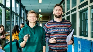 Twee witte mannen, Nicolaas Veul links en Tim den Besten rechts, lopen door een schoolgang; de een draagt een groene hoodie en de ander een kleurrijke trui en houdt papieren vast. Op de achtergrond zitten vier jonge mensen op een bank, onder wie een zwarte vrouw in een gele trui.