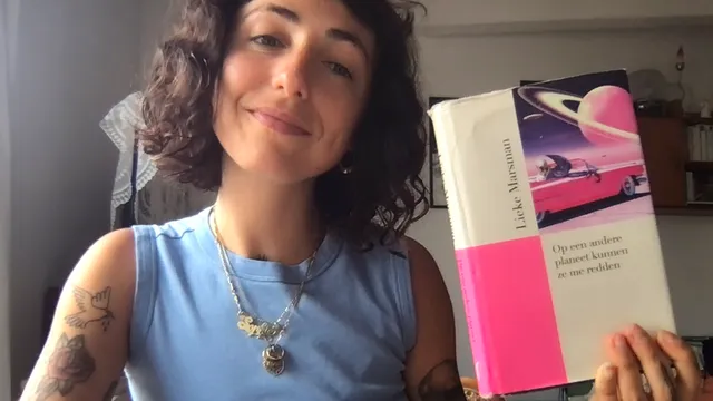 Sophie Straat glimlacht in de camera en houdt het boek Op een andere planeet kunnen ze me redden van Lieke Marsman naast haar hoofd vast.