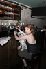 Een persoon met kort, roodbruin haar zit op een kruk aan een bar en kijkt omhoog naar de camera. Ze dragen een zwarte top met een open rug, waarop een tatoeage te zien is. Op hun schoot zit een kleine, harige, wit-grijze hond die recht in de lens kijkt. Met één hand houdt de persoon een glas vast terwijl ze met de andere hand een drankje inschenken uit een flesje. De bar heeft een achterwand van bakstenen en een stellingkast vol glazen en flessen. Op de tapknoppen op de voorgrond is het logo van Gulpener te zien.