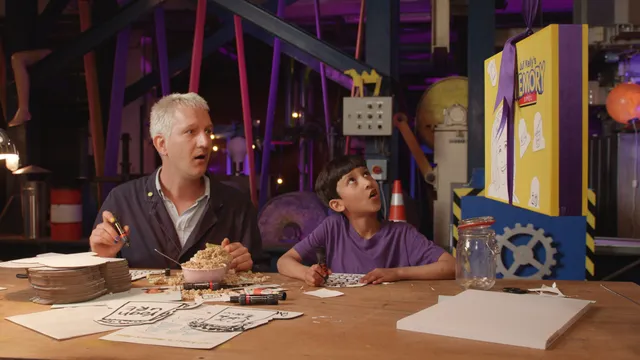 Presentator Maarten Bel zit samen met een kind aan een knutseltafel. Ze kijken allebei naar de zijkant met hun mond geopend.