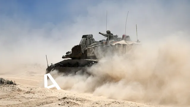 Een tank rijdt door een stoffig woestijnlandschap, waarbij een soldaat uit de luikopening bovenop het voertuig een gebaar maakt. Het zicht wordt deels belemmerd door opwaaiend stof.