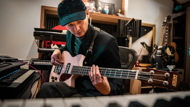 Een man achter een laptop in een muziekstudio met een roze basgitaar op schoot