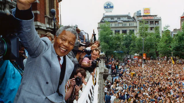Nelson Mandela staat op het balkon van de Stadsschouwburg op het Leidseplein in Amsterdam. Hij heft zijn vuist als groet naar de grote mensen massa en fotografen.