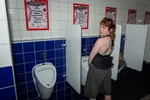Een persoon met kort roodbruin haar en een zonnebril op het hoofd staat omgedraaid bij een urinoir in een openbaar toilet en kijkt achterom naar de camera. De persoon draagt een zwarte, mouwloze top met ruches aan de achterkant en een wijde, donkergrijze korte broek. De toiletruimte heeft witte tegels aan de bovenkant en blauwe tegels aan de onderkant, gescheiden door witte schotten tussen de urinoirs. Aan de muur boven elk urinoir hangt een rode poster met de tekst 'Daddyfest'.