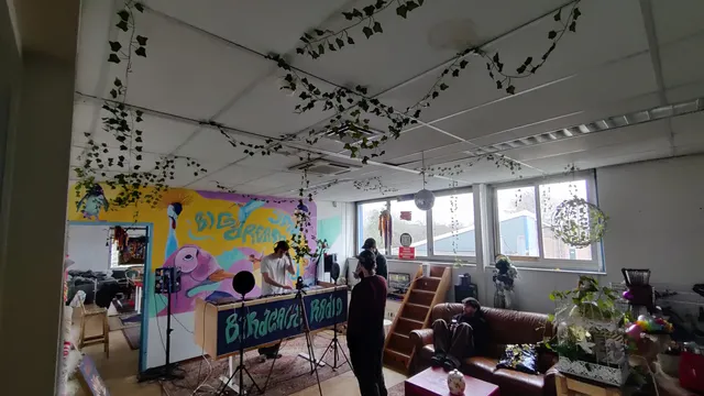 Een sfeervolle overzichtsfoto van een creatieve binnenruimte waar 'Birdcage Radio' live uitzendt. Een dj in een wit shirt staat achter een mengpaneel dat op een houten meubel is geplaatst, met daarop de tekst 'Birdcage Radio'. Hij draagt een koptelefoon en wordt gefilmd door een camera op statief. Op de achtergrond is een kleurrijke wandschildering te zien met tropische vogels en de tekst 'BIG DREAM'. In de leefruimte staan mensen te kijken en zit er iemand op een leren bank. Het systeemplafond is versierd met hangende kunstklimop. De ruimte is gevuld met kunst, planten en persoonlijke items.
