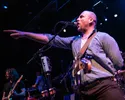 Foto van de frontman van Iguana Death Cult die in de microfoon zingt en de zaal in wijst met een gestrekte arm.