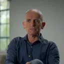 Niels van Elk zit ontspannen in een stoel in een lichte woonkamer met hoge ramen. Hij draagt een blauw linnen overhemd en jeans, en kijkt rustig en direct in de camera.