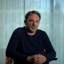 Jochem Pinxteren zit in een stoel in een lichte ruimte met verticale gordijnen. Hij draagt een donkerblauwe trui, heeft zijn handen gevouwen en kijkt serieus opzij.