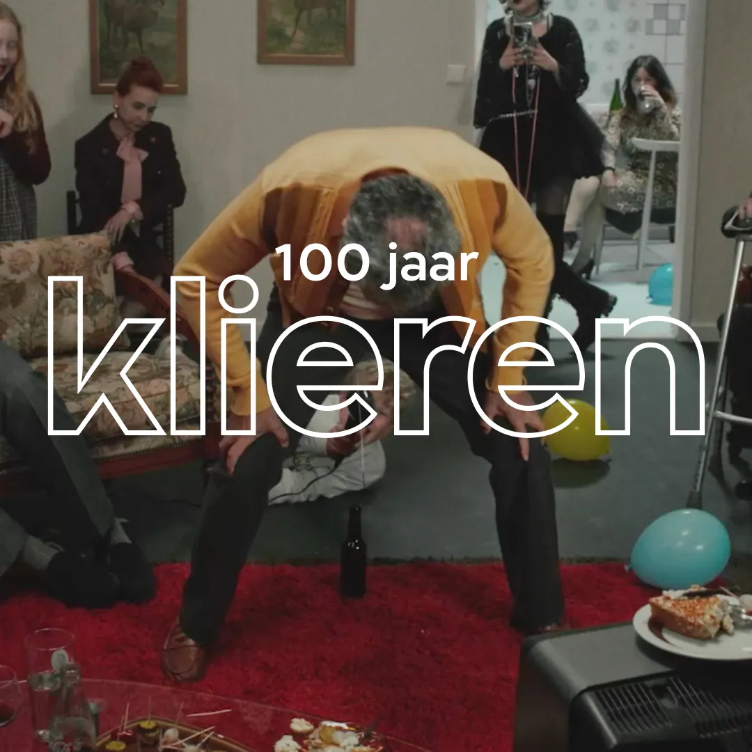 Man die in een woonkamer aan het spijkerpoepen is. De tekst 100 jaar klieren in beeld.