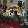Man die in een woonkamer aan het spijkerpoepen is. De tekst 100 jaar klieren in beeld.