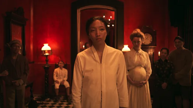 Vrouw in licht gewaad staat vooraan in rood verlichte kamer met zwart-wit vloer en enkele mensen op de achtergrond.