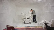 Een kunstenaar werkt in een volledig witte studio aan een sculptuur van paarden. Naast hem staat een zwarte hond, en op de vloer liggen meer papier-maché vormen.