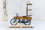 Een blauwe bromfiets met een houten bekisting staat vastgesjord op een houten pallet in een lichte expositieruimte. Achterop is een hoge, doorzichtige cilindervormige installatie gemonteerd, met daarin een geelachtig hangend object. Oranje slangen verbinden de installatie met de bromfiets. De achtergrond bestaat uit een witte muur en een betonnen vloer.