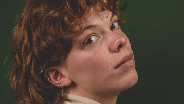 Een medium close-up studioportret van Maxime met rood, krullend haar in een matje (mullet). Maxime draagt een witte coltrui, een crèmekleurig jack met stiksels, subtiele oorbellen en een neuspiercing, en kijkt met een neutrale blik over de schouder naar de camera tegen een donkergroene achtergrond.
