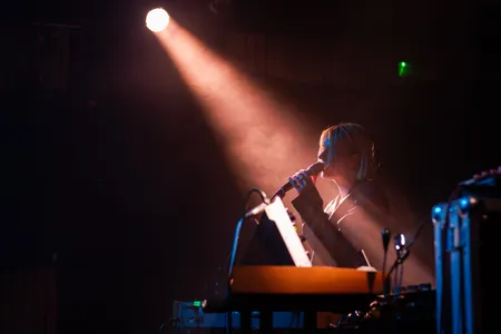 Een sfeervol beeld van een zangeres op een donker podium, verlicht door een enkele felle lichtbundel van bovenaf. Ze staat zijdelings en houdt een microfoon dicht bij haar mond. Op de voorgrond zijn de contouren van een keyboard en andere geluidsapparatuur zichtbaar in een warme, oranje gloed, terwijl er een lichte nevel door het licht danst.