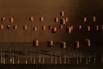 Een kunstinstallatie van tientallen kleine, roestbruine cilindervormige objecten op lange, dunne metalen staven. De cilinders staan op verschillende hoogtes en lijken boven de grond te zweven. De donkere ruimte is verlicht met warm oranjebruin licht, waardoor lange schaduwen ontstaan.