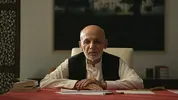 Een oudere man met een lichtbruine huid, kaal hoofd en grijze baard zit achter een tafel met papieren en boeken. Hij draagt een wit gewaad met een zwart vest en kijkt recht in de camera.
