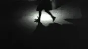Zwart-witbeeld van een nachtelijke scène waarin een persoon in een rok of jurk over een betegeld oppervlak loopt. Alleen de benen zijn zichtbaar in een kegel van licht; de rest van het lichaam gaat op in de schaduw. De persoon werpt een lange schaduw vooruit.