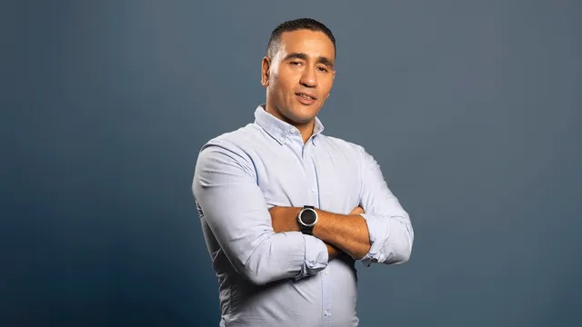 Abdou Bouzerda, gekleed in een lichtblauw overhemd en zwarte broek, staat met gekruiste armen tegen een neutrale grijsblauwe achtergrond en kijkt zelfverzekerd in de camera.