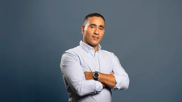 Abdou Bouzerda, gekleed in een lichtblauw overhemd en zwarte broek, staat met gekruiste armen tegen een neutrale grijsblauwe achtergrond en kijkt zelfverzekerd in de camera.