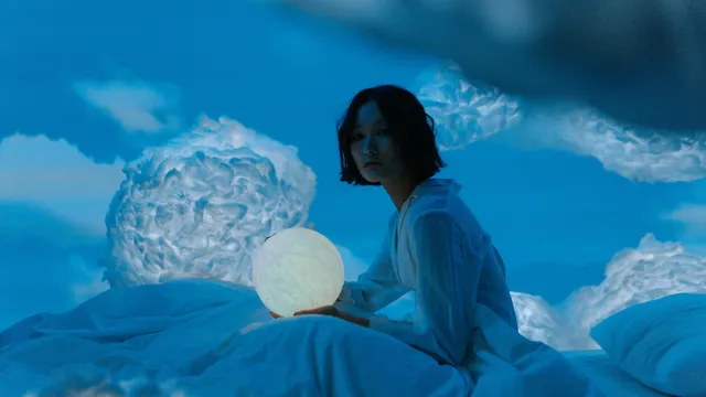 Vrouw zit rechtop onder witte lakens, omringd met wolken en heeft een lichtgevende bol in haar handen.