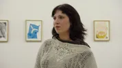 Een vrouw met donker haar en een lichte gebreide trui staat in een witte tentoonstellingsruimte. Achter haar hangen kleurrijke schilderijen aan de muur.