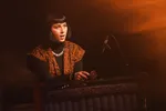 Een vrouw met een zwarte bob en een opvallende zilveren ketting staat achter een houten marimba of xylofoon met houtsnijwerk. Ze heeft haar mond open alsof ze zingt of spreekt. De scène is sfeervol verlicht met een warme oranje gloed en er is een microfoon zichtbaar.
