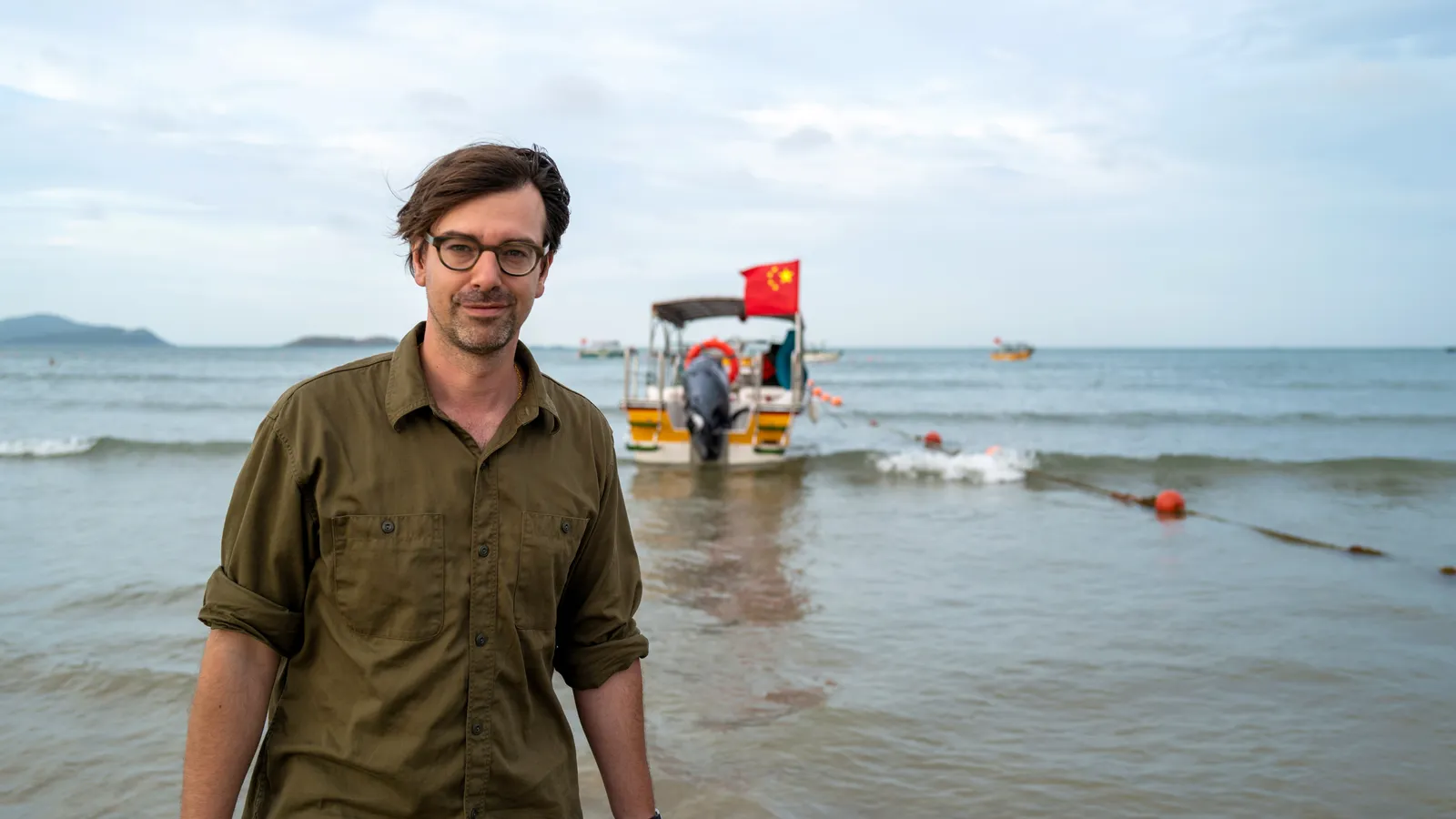Een witte man met donkerbruin haar en een bril staat in ondiep water op het strand, met een legergroen overhemd aan. Achter hem ligt een boot met een Chinese vlag, aangemeerd in zee. Op de achtergrond zijn meerdere boten en een heuvelachtig landschap zichtbaar.
