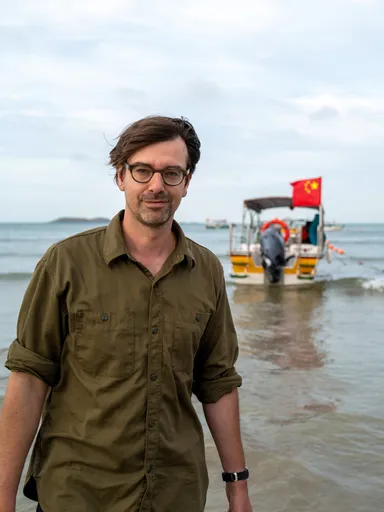 Een witte man met donkerbruin haar en een bril staat in ondiep water op het strand, met een legergroen overhemd aan. Achter hem ligt een boot met een Chinese vlag, aangemeerd in zee. Op de achtergrond zijn meerdere boten en een heuvelachtig landschap zichtbaar.