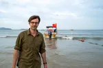 Een witte man met donkerbruin haar en een bril staat in ondiep water op het strand, met een legergroen overhemd aan. Achter hem ligt een boot met een Chinese vlag, aangemeerd in zee. Op de achtergrond zijn meerdere boten en een heuvelachtig landschap zichtbaar.