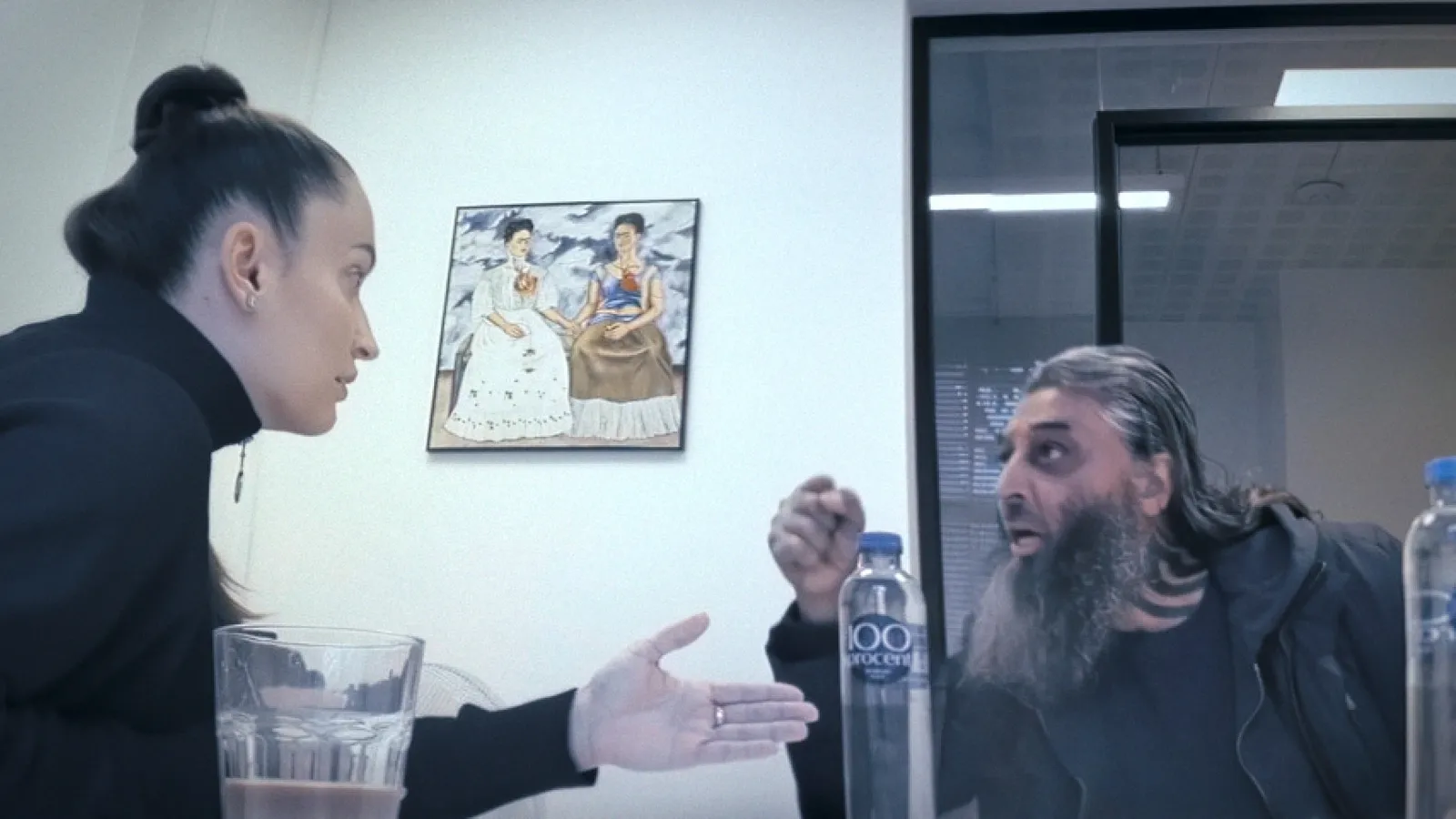 Twee mensen in gesprek aan een tafel in een kantoorruimte: een witte vrouw met strak naar achter gebonden donker haar en een oorbel gebaart naar een man met een donkere huidskleur, lange grijze baard en lang haar, die nadrukkelijk iets zegt. Op tafel staan glazen en flessen water. Aan de muur hangt een schilderij van twee vrouwen.